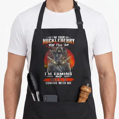Huckleberry novelty apron - Skeleton cowboy in red circle with text I'm Your Huckleberry You Tell Em I'm Coming — shown on a black cotton canvas apron against a white background