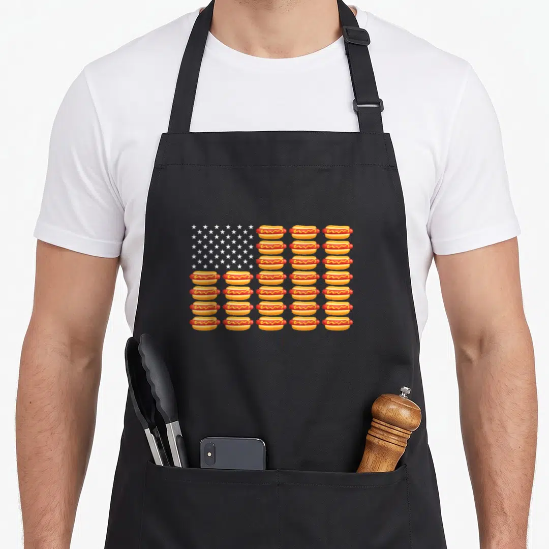 Flag hot dog apron - Cartoon USA flag formed by rows of hot dogs with white stars in blue corner — shown on a black cotton canvas apron against a white background