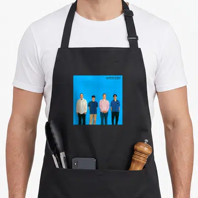 Weezer cover apron - Four band members standing against blue background with black lowercase text in corner — shown on a black cotton canvas apron against a white background