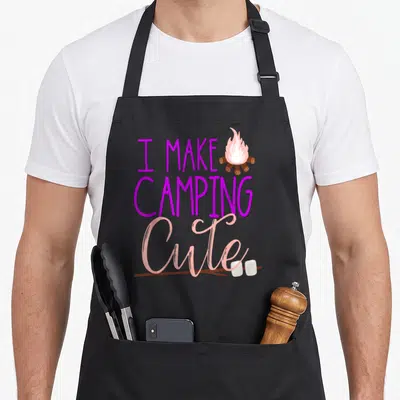 Daughter camping apron - Purple text I Make Camping Cute with cartoon campfire and marshmallow stick — shown on a black cotton canvas apron against a white background