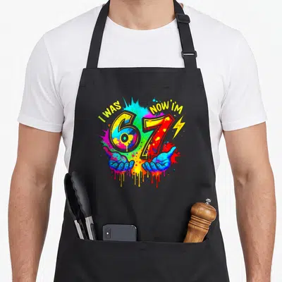 Six paint apron - I WAS NOW I'M 67 with two hands and lightning bolt on paint splatter background — shown on a black cotton canvas apron against a white background