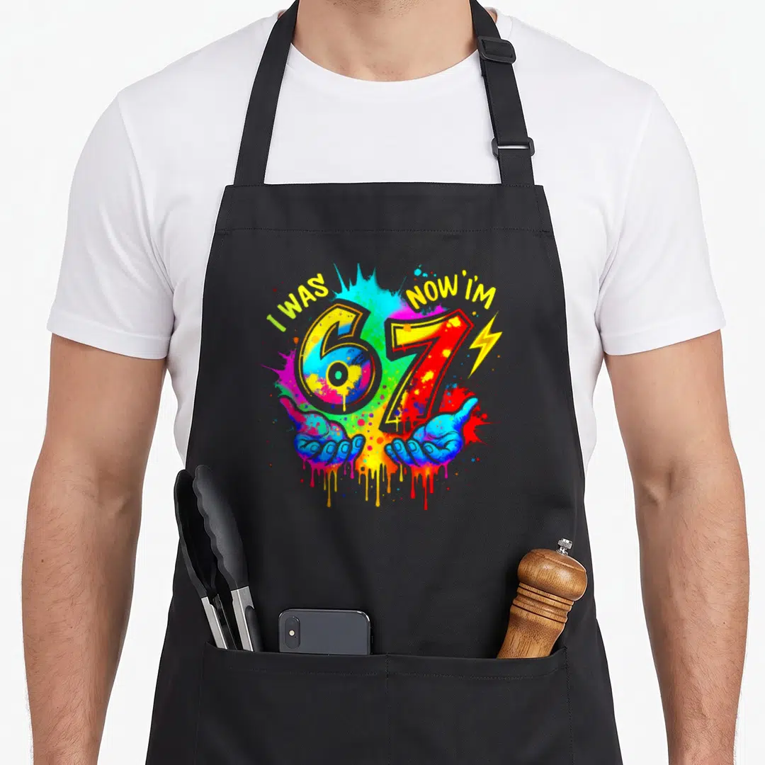 Six paint apron - I WAS NOW I'M 67 with two hands and lightning bolt on paint splatter background — shown on a black cotton canvas apron against a white background