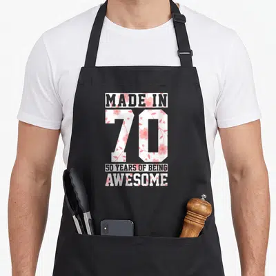 Funny 50th vintage floral apron - Stacked text Made In 70 50 Years Of Being Awesome with pink floral pattern fill — shown on a black cotton canvas apron against a white background