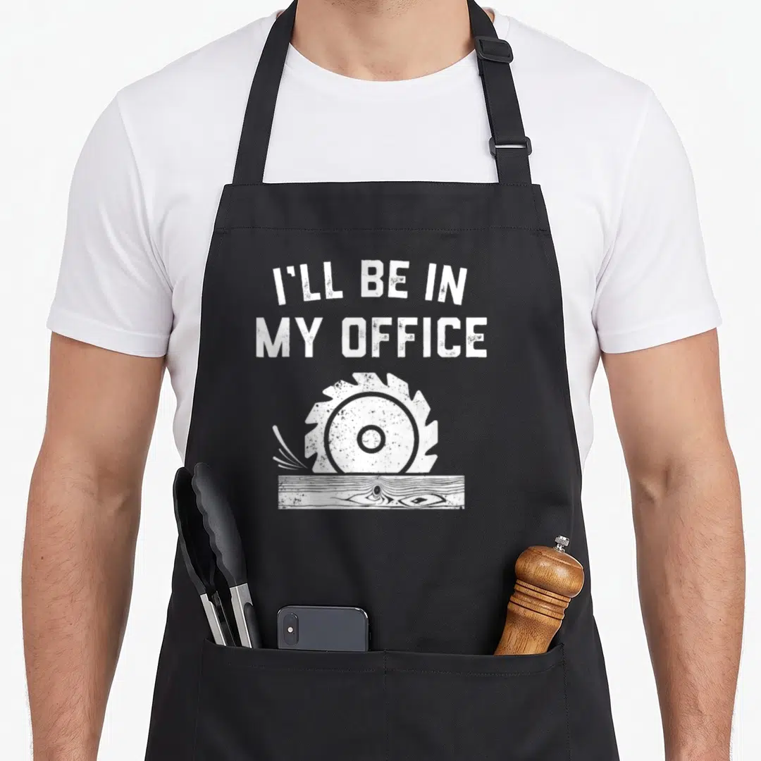 Woodworking father handyman apron - White distressed circular saw cutting wood plank below stacked text I'll be in my — shown on a black cotton canvas apron against a white background