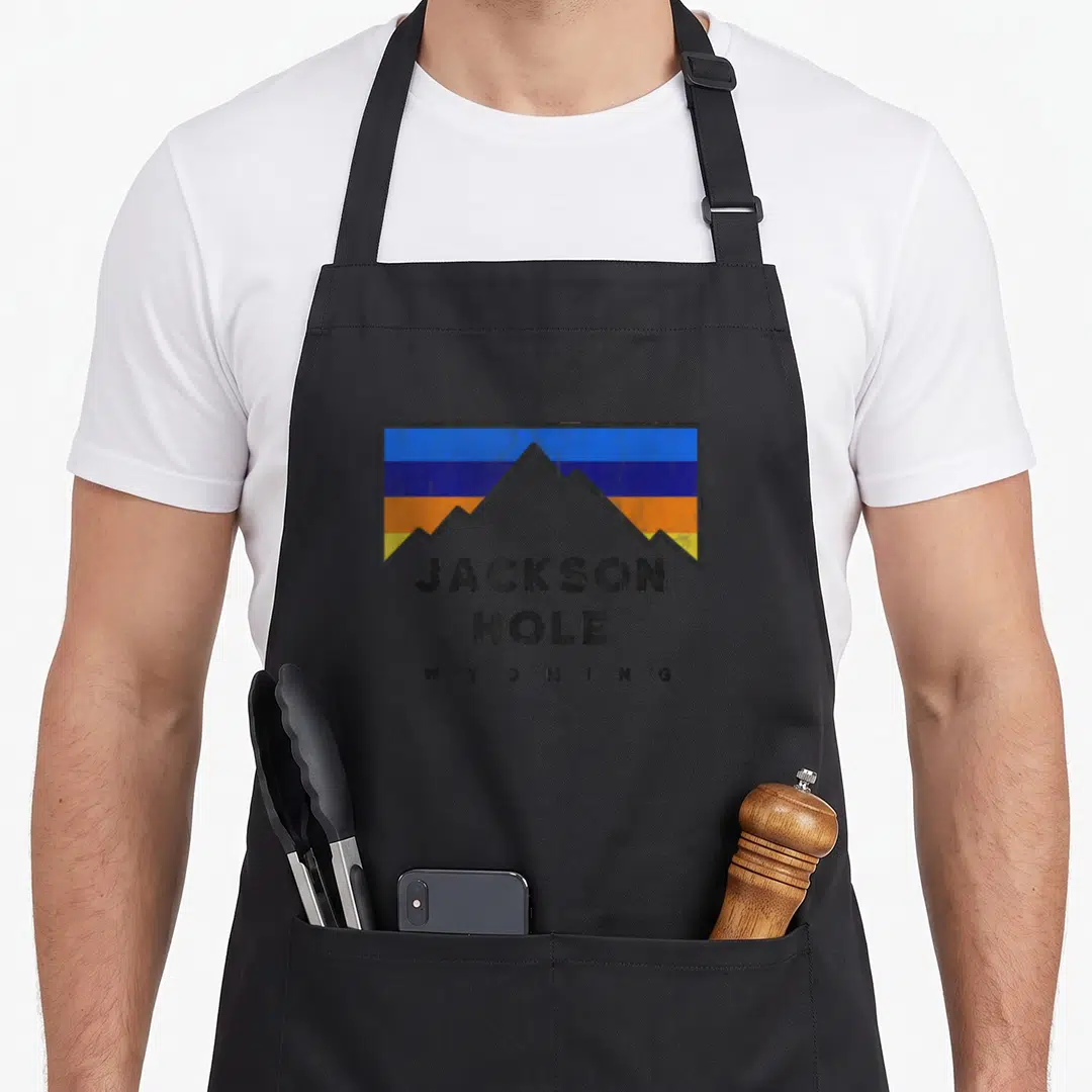 Hole jackson apron - Retro mountain silhouette with blue orange yellow grey stripes and stacked JACKSON HOLE WYOMING — shown on a black cotton canvas apron against a white background