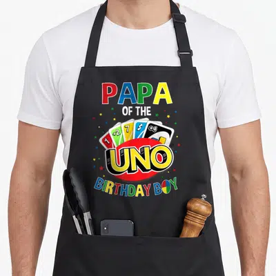 Uno family aprons - Papa of the Uno Birthday Boy text with fanned playing cards and colorful stars — shown on a black cotton canvas apron against a white background