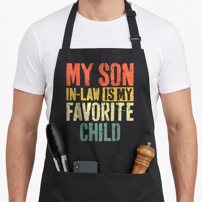 Law mother father day apron - Distressed text MY SON IN-LAW IS MY FAVORITE CHILD in orange yellow beige and green — shown on a black cotton canvas apron against a white background