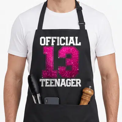 Bday tween birthday apron - OFFICIAL 13 TEENAGER in pink glitter and white block letters stacked vertically — shown on a black cotton canvas apron against a white background