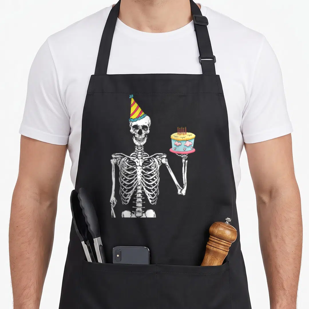 Lazy funny halloween apron - White cartoon skeleton wearing striped party hat holding cake with blue frosting and pink — shown on a black cotton canvas apron against a white background