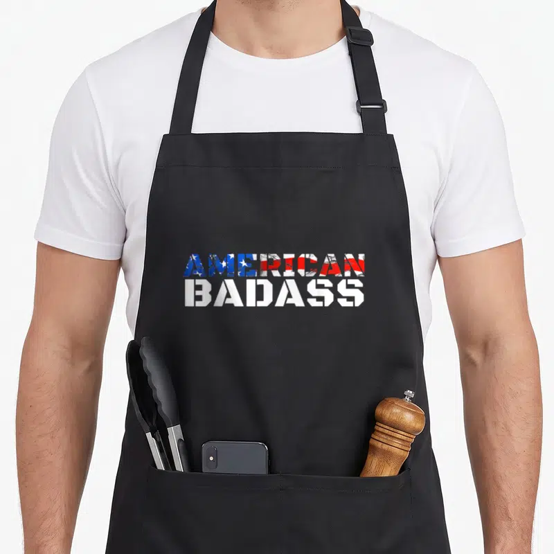 Flag badass apron - AMERICAN BADASS text with USA flag pattern fill and solid white letters — shown on a black cotton canvas apron against a white background