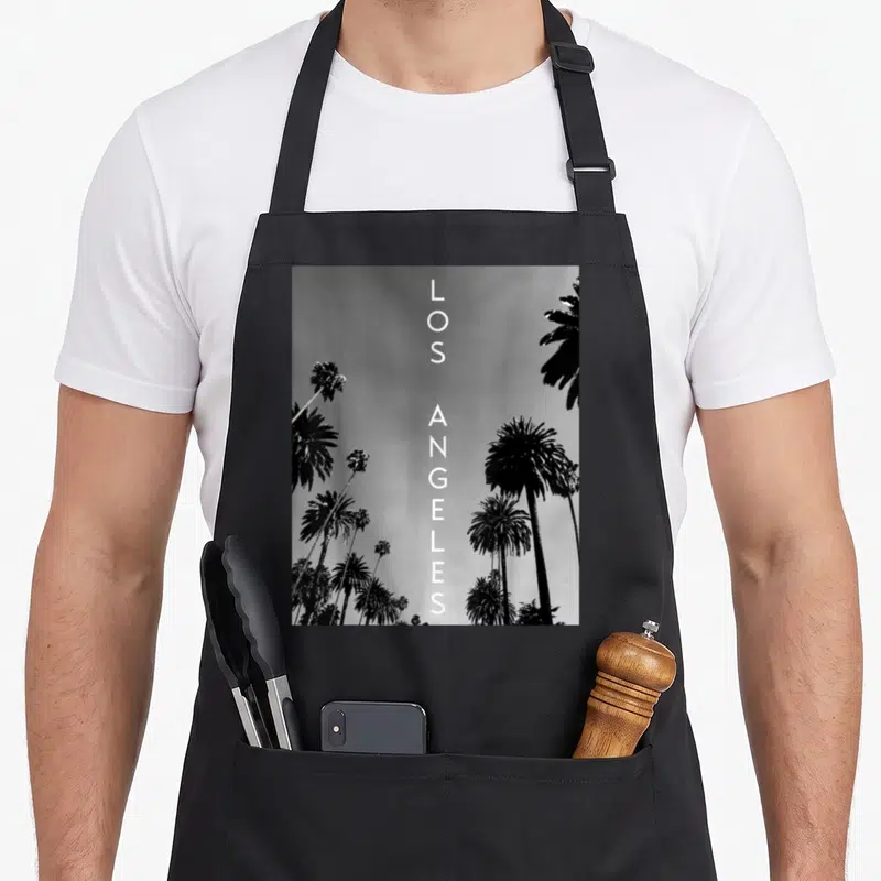 Los laos apron - Vertical LOS ANGELES text flanked by black and white palm trees against a grey sky — shown on a black cotton canvas apron against a white background