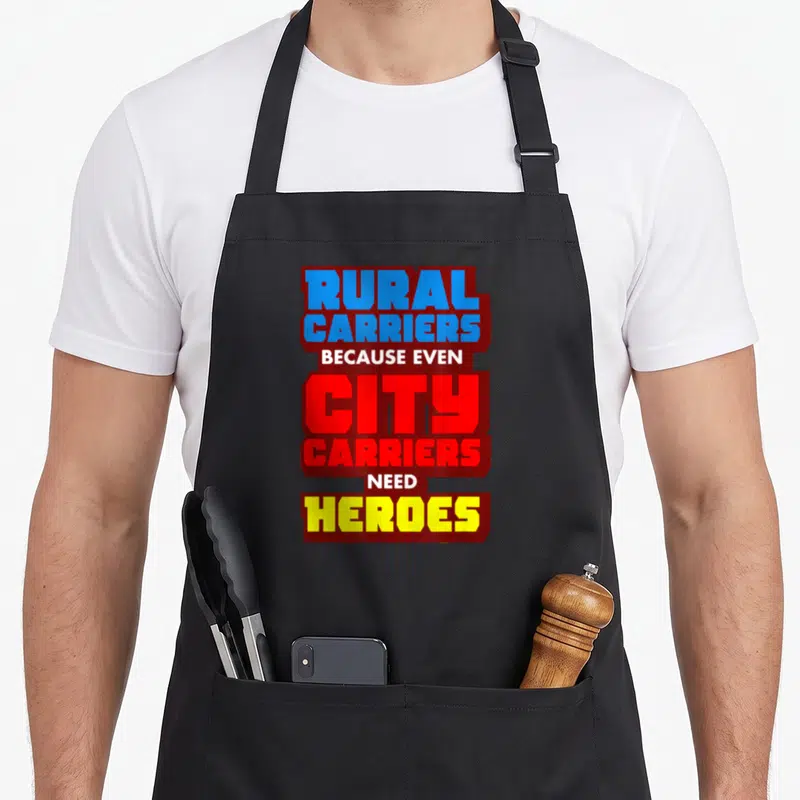 Carrier postman postal aprons - Stacked text reading RURAL CARRIERS BECAUSE EVEN CITY CARRIERS NEED HEROES in blue red — shown on a black cotton canvas apron against a white background