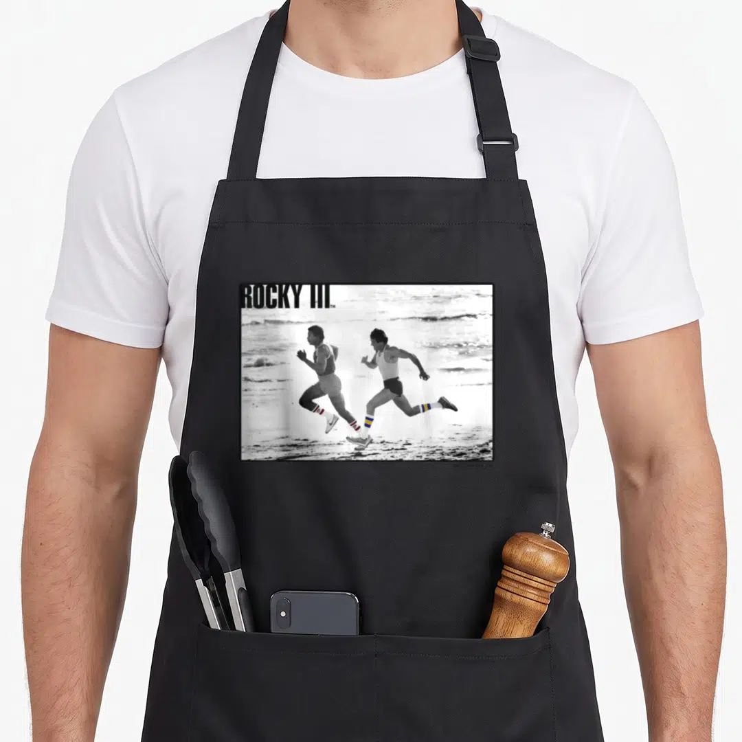Rocky apollo apron - Two men running on a beach in a black and white photo with ROCKY III text overlay — shown on a black cotton canvas apron against a white background