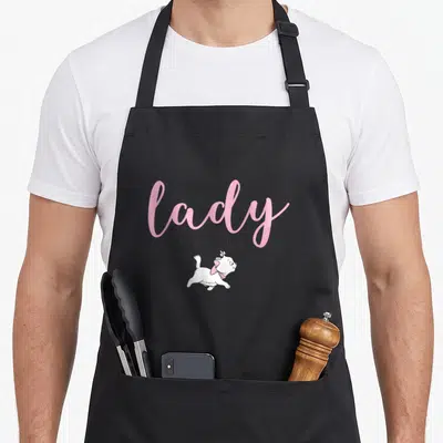 Marie novelty apron - Stacked text Because I'm A Lady above a running white cartoon cat with a pink bow on grey — shown on a black cotton canvas apron against a white background