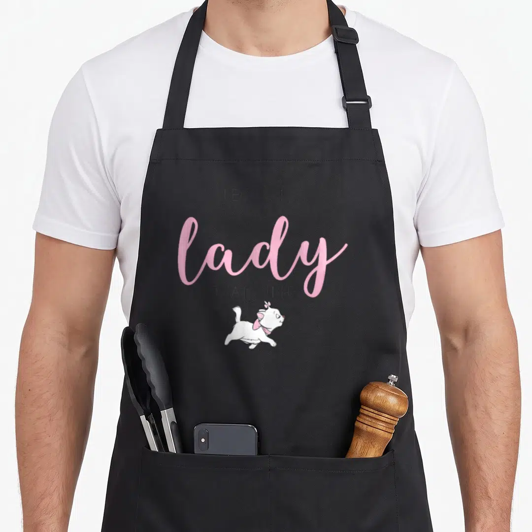 Marie novelty apron - Stacked text Because I'm A Lady above a running white cartoon cat with a pink bow on grey — shown on a black cotton canvas apron against a white background