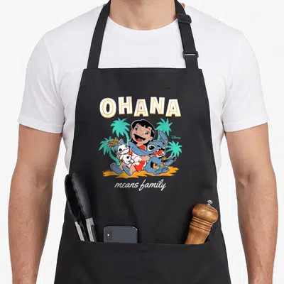 Ohana mean family aprons - Lilo holding doll and Stitch with guitar under OHANA text on orange sand — shown on a black cotton canvas apron against a white background