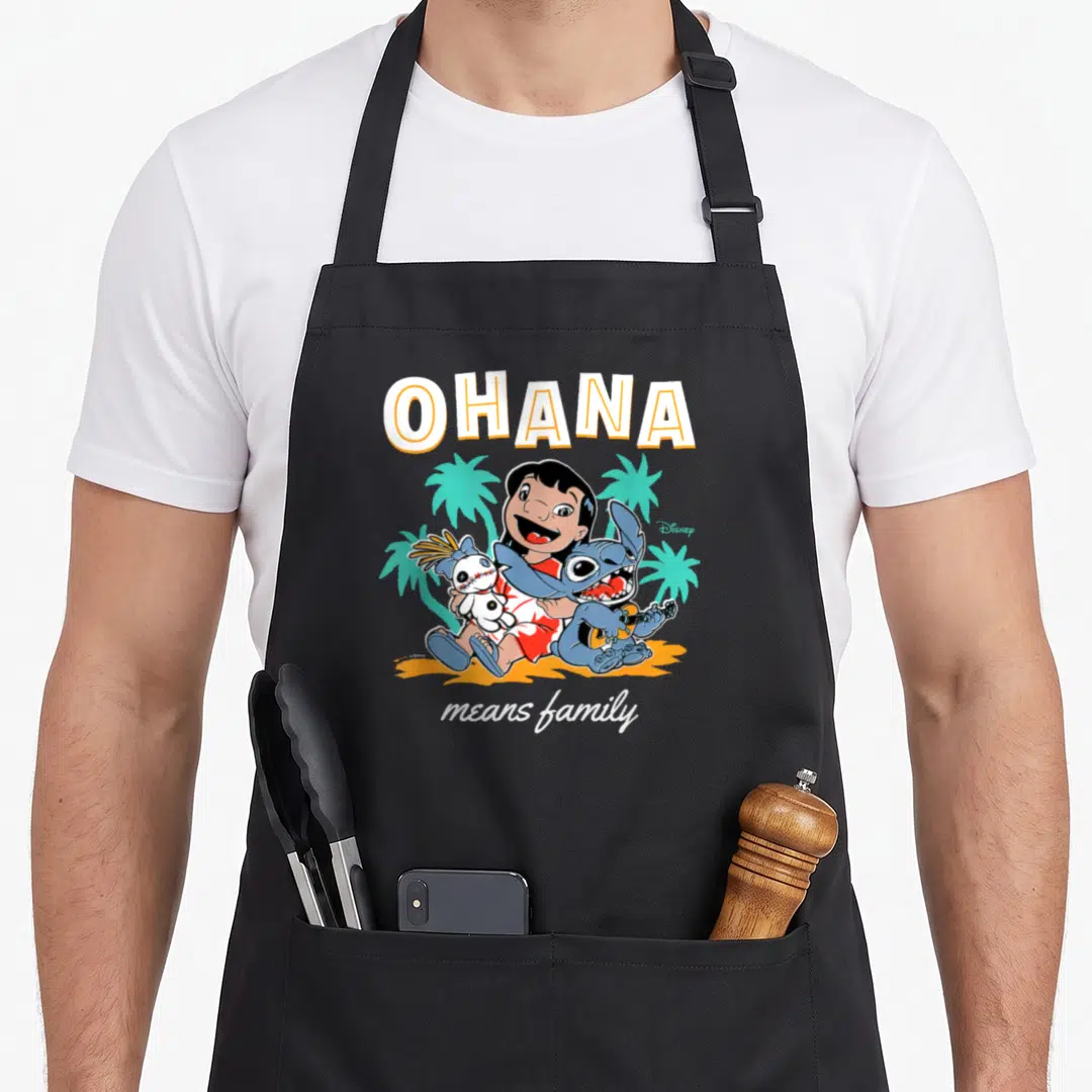 Ohana mean family aprons - Lilo holding doll and Stitch with guitar under OHANA text on orange sand — shown on a black cotton canvas apron against a white background