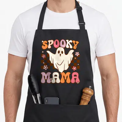 Ghost mama apron - Cartoon ghost over brown checkerboard with SPOOKY MAMA text in pink and orange — shown on a black cotton canvas apron against a white background