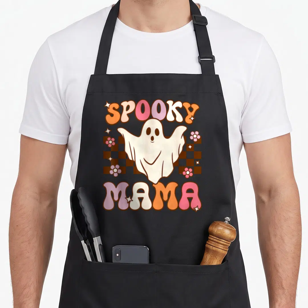 Ghost mama apron - Cartoon ghost over brown checkerboard with SPOOKY MAMA text in pink and orange — shown on a black cotton canvas apron against a white background