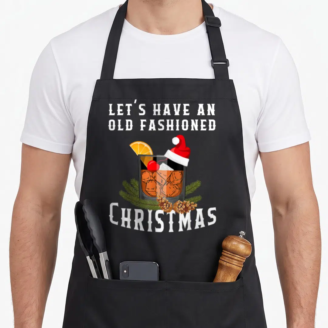 Drink old fashioned aprons - Old Fashioned cocktail with Santa hat, pine branches, and festive text — shown on a black cotton canvas apron against a white background