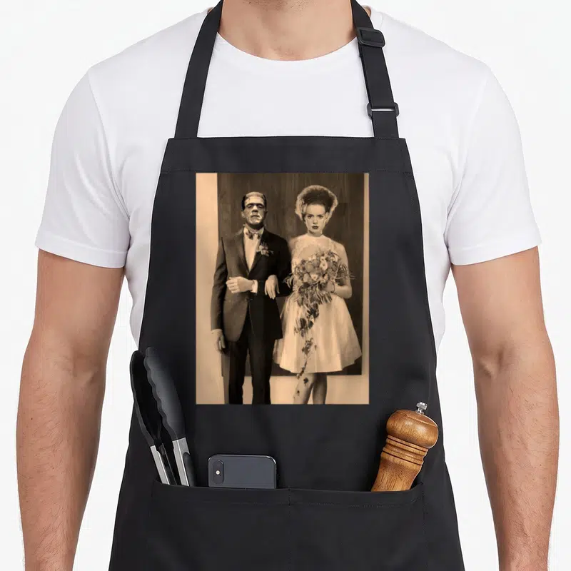 Dapper wedding day aprons - Sepia photo of Frankenstein groom and Bride of Frankenstein holding bouquet against wood — shown on a black cotton canvas apron against a white background