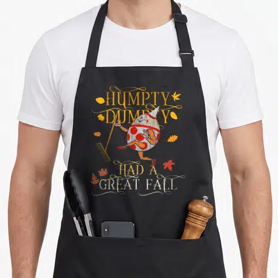 Funny humpty cute fall aprons - Humpty Dumpty holding a rake surrounded by yellow and red autumn leaves with distressed — shown on a black cotton canvas apron against a white background