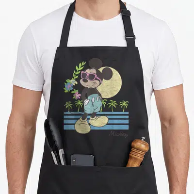 Beach friends apron - Retro Mickey Mouse in pink sunglasses with palm trees, flowers, and yellow sun — shown on a black cotton canvas apron against a white background