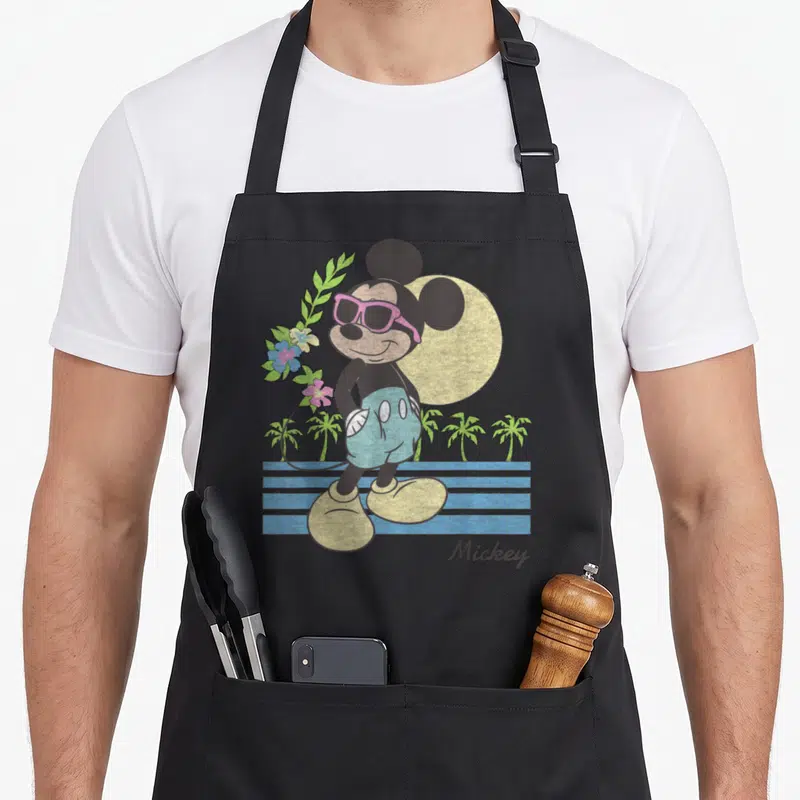 Beach friends apron - Retro Mickey Mouse in pink sunglasses with palm trees, flowers, and yellow sun — shown on a black cotton canvas apron against a white background