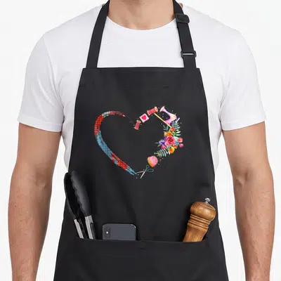 Quilting sewing needlework apron - Watercolor heart shape formed by sewing tools and flowers in pink red blue and green — shown on a black cotton canvas apron against a white background