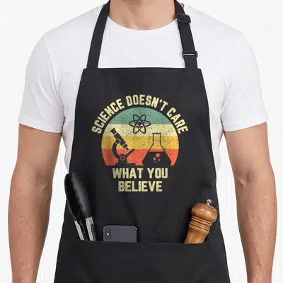 Care science apron - Retro striped circle with atom, microscope, flask and text SCIENCE DOESN'T CARE WHAT YOU BELIEVE — shown on a black cotton canvas apron against a white background