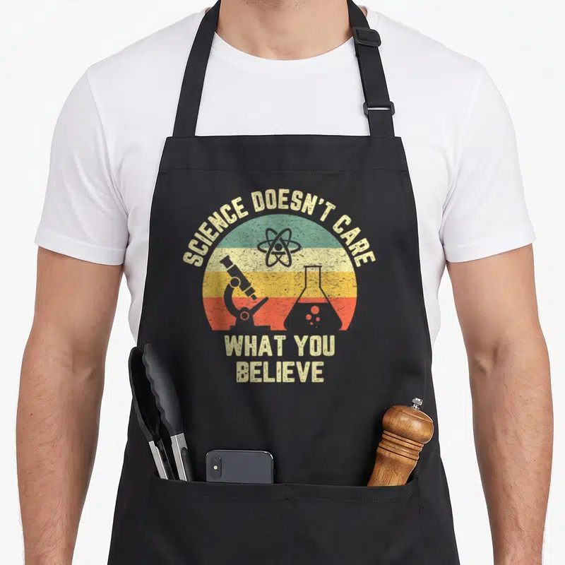 Care science apron - Retro striped circle with atom, microscope, flask and text SCIENCE DOESN'T CARE WHAT YOU BELIEVE — shown on a black cotton canvas apron against a white background