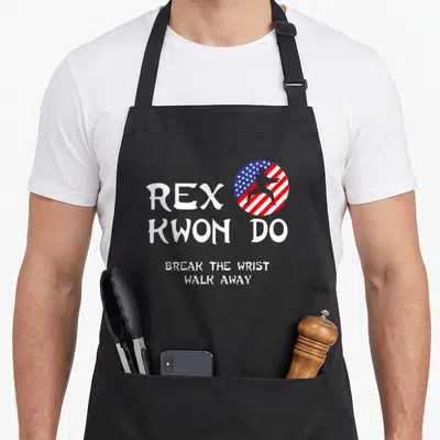 Break t-rex apron - REX KWON DO text and martial artist silhouette in red white blue circle — shown on a black cotton canvas apron against a white background