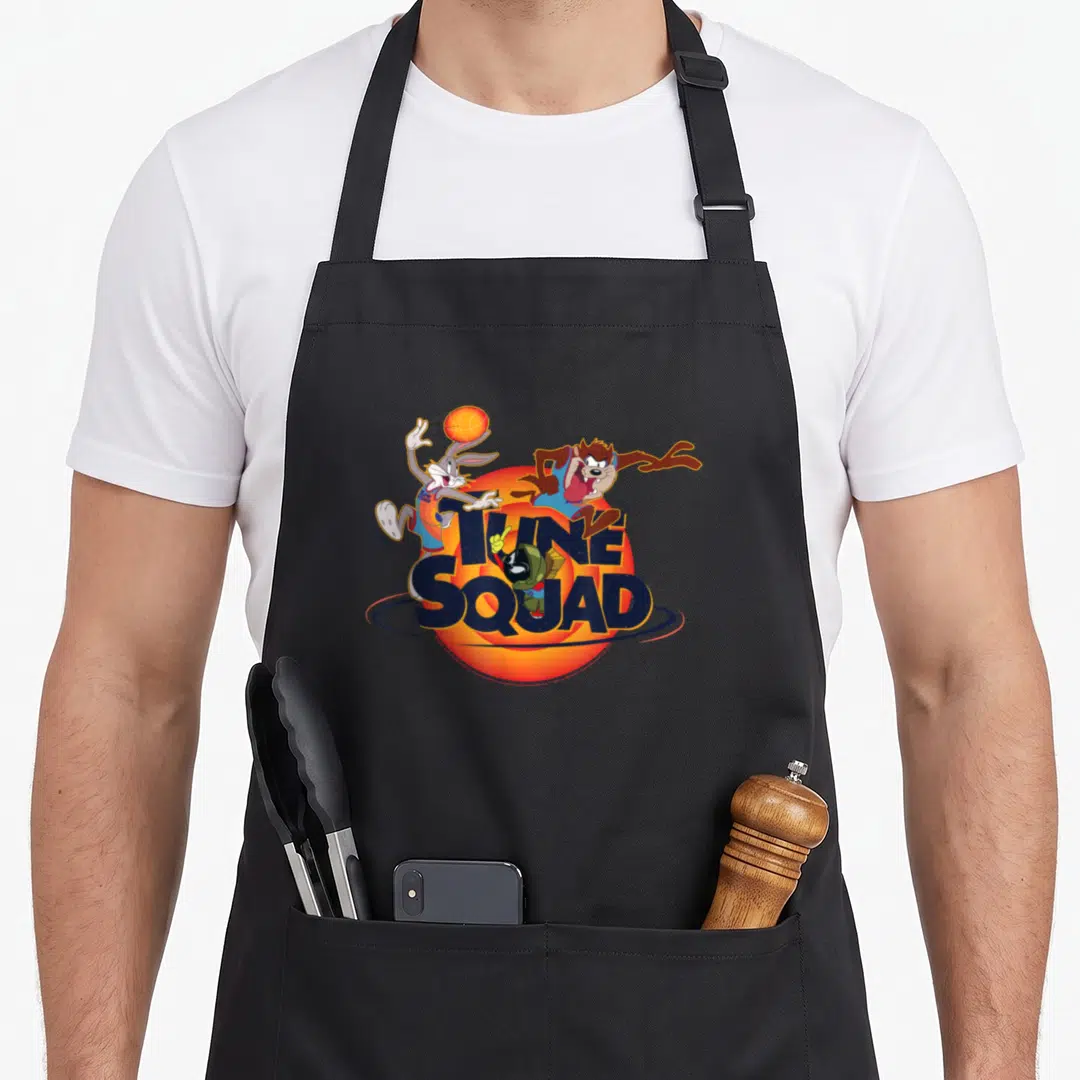 Bug jam apron - Tune Squad text with Bugs Bunny, Tasmanian Devil, and Marvin the Martian over orange circle — shown on a black cotton canvas apron against a white background