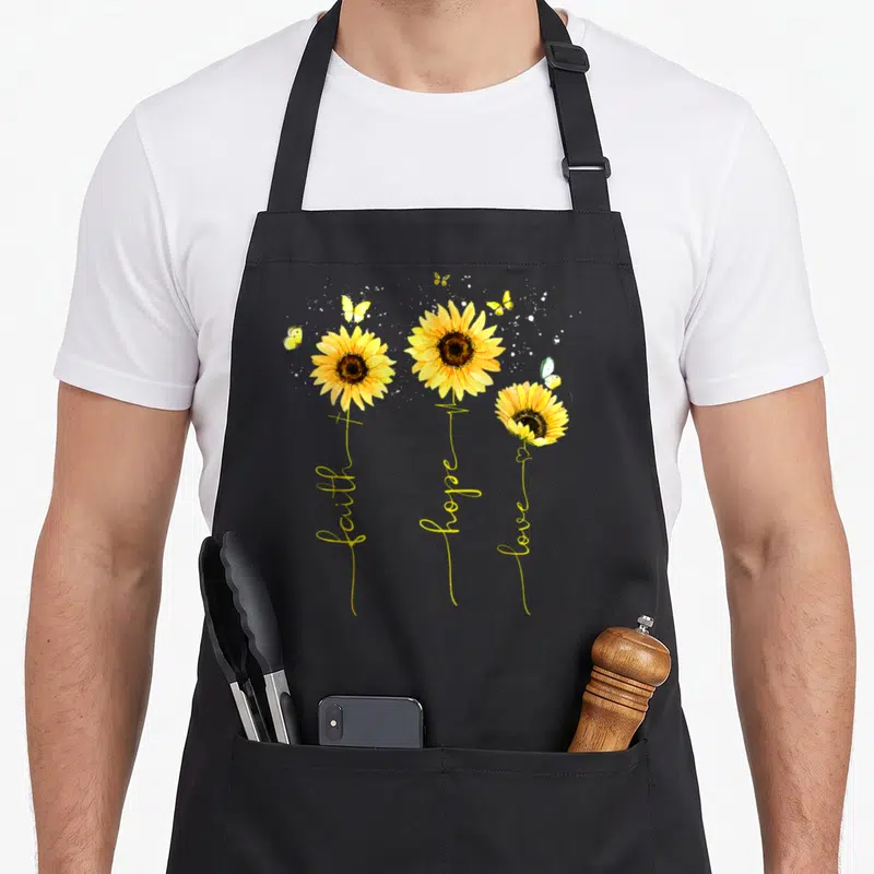Christian sunflower apron - Three yellow sunflowers with stems forming faith hope love text, white splatters — shown on a black cotton canvas apron against a white background
