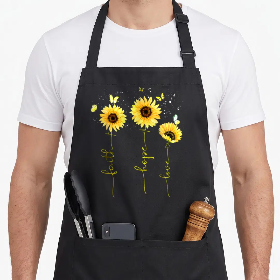Christian sunflower apron - Three yellow sunflowers with stems forming faith hope love text, white splatters — shown on a black cotton canvas apron against a white background