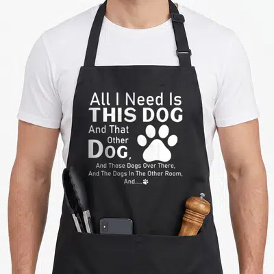 Those dog apron - White stacked text reading All I Need Is This Dog with a large paw print on the right side — shown on a black cotton canvas apron against a white background