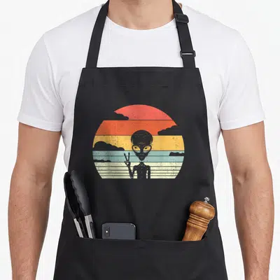 Extraterrestrial alien apron - Grey alien holding up two fingers in front of a multi-colored striped sun — shown on a black cotton canvas apron against a white background