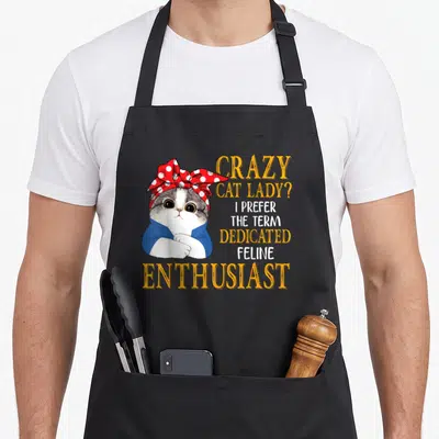 Prefer crazy cat lady apron - Cartoon grey and white cat wearing a red polka dot bow and blue shirt next to orange — shown on a black cotton canvas apron against a white background