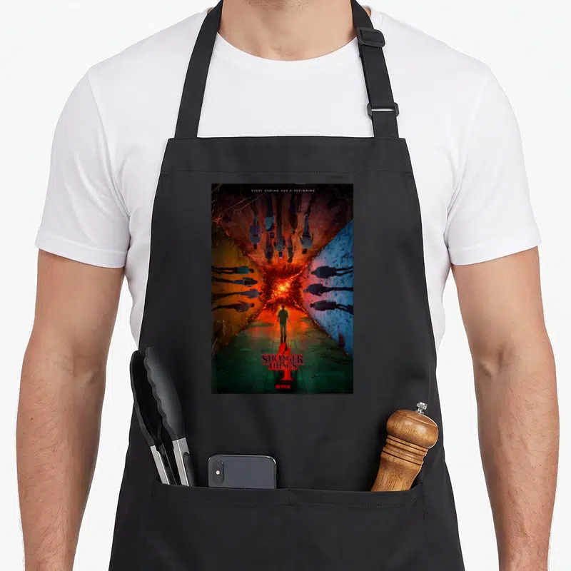 Hallway novelty apron - Man facing red portal in corridor with Stranger Things 4 logo and quote — shown on a black cotton canvas apron against a white background