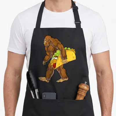 Bigfoot taco apron - Cartoon brown bigfoot holding giant yellow taco with green lettuce and fries — shown on a black cotton canvas apron against a white background
