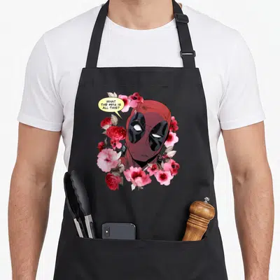 Flower crown novelty apron - Deadpool head surrounded by red and pink flowers with a yellow speech bubble containing — shown on a black cotton canvas apron against a white background