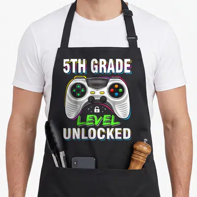 5th level apron - Game controller graphic with 5th Grade Level Unlocked text and small lock icon — shown on a black cotton canvas apron against a white background