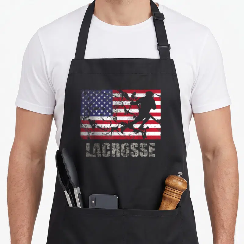 Lacrosse fan american flag apron - Distressed USA flag with grey lacrosse player silhouette and text below — shown on a black cotton canvas apron against a white background