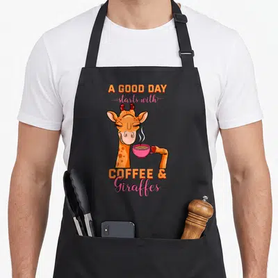 Giraffe coffee bean apron - Cartoon giraffe holding pink cup with A Good Day starts with Coffee and Giraffes text — shown on a black cotton canvas apron against a white background