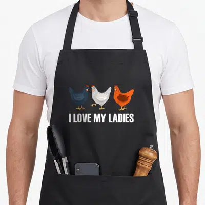 Coop chicken with apron - Three cartoon hens in black, white, orange, and red above I LOVE MY LADIES text — shown on a black cotton canvas apron against a white background