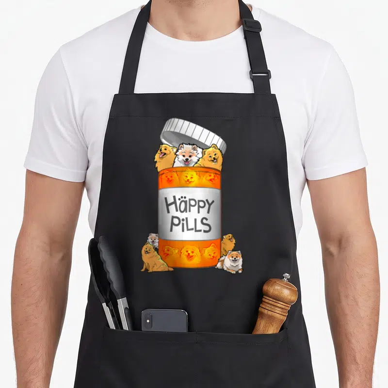 Pill pomeranian apron - Orange pill bottle with cartoon Pomeranians and Häppy PiLLS text on black fabric — shown on a black cotton canvas apron against a white background