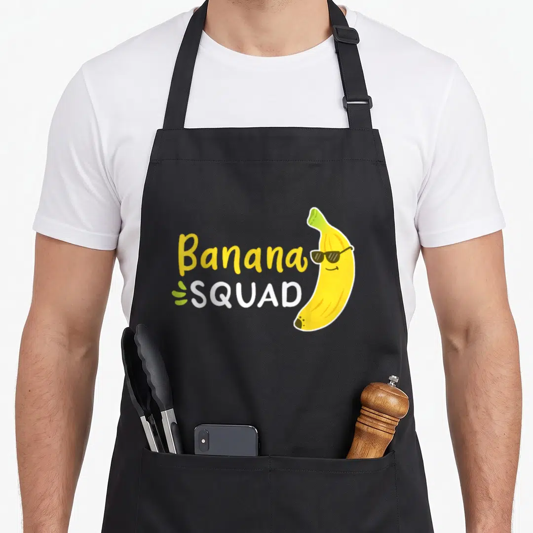 Banana fruit apron - Cartoon banana in sunglasses beside stacked Banana SQUAD text with green leaf accent — shown on a black cotton canvas apron against a white background