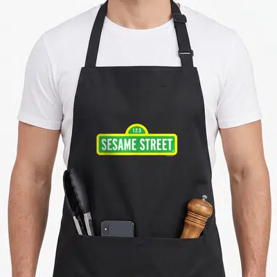 Cute street apron - Green and yellow Sesame Street sign with 123 and white text — shown on a black cotton canvas apron against a white background