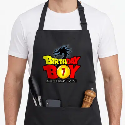 7th anime mom apron - Black dragon head above BIRTHDAY BOY text with yellow 7 sphere and Japanese greeting — shown on a black cotton canvas apron against a white background
