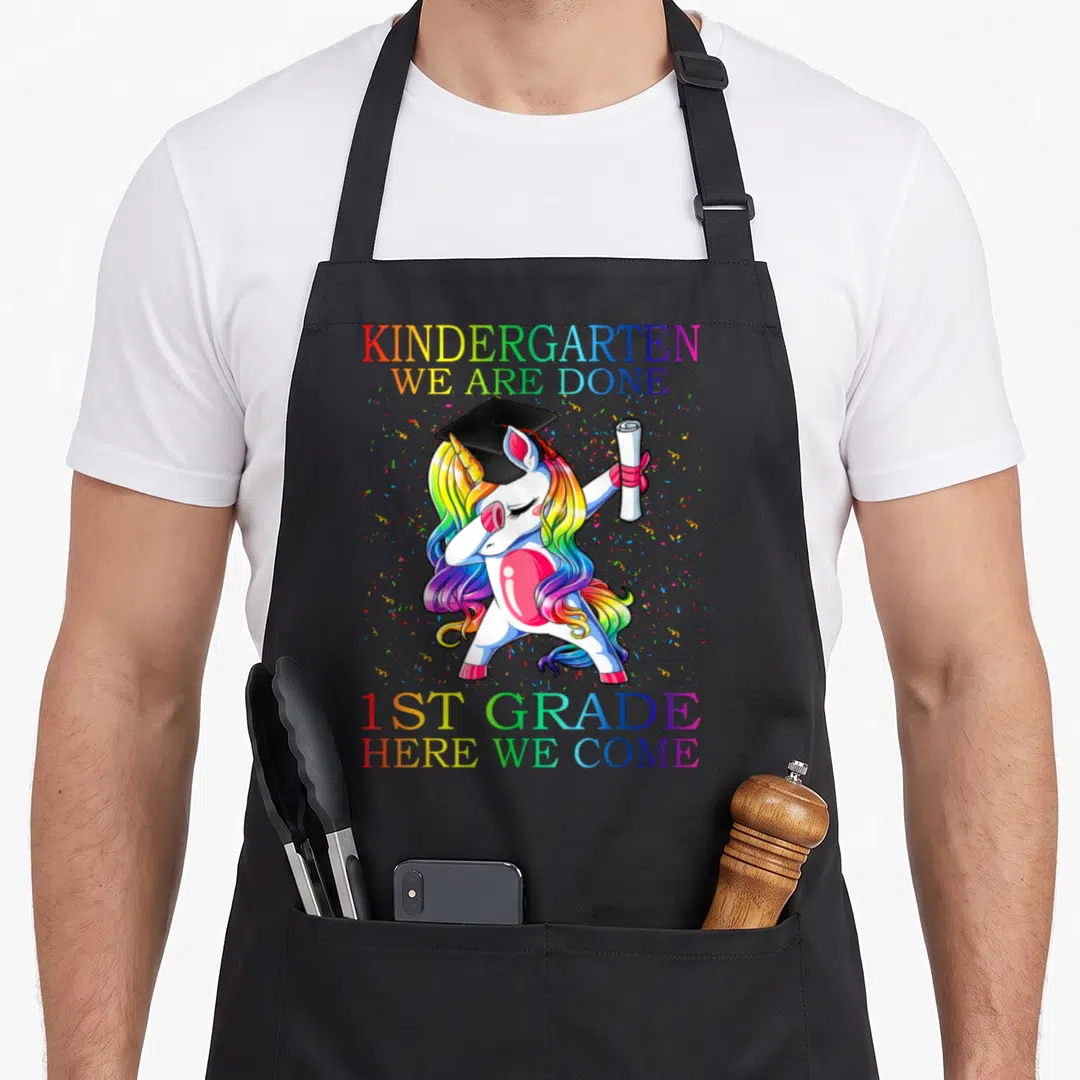 1st unicorn aprons - White unicorn dabbing with cap and diploma between rainbow text blocks — shown on a black cotton canvas apron against a white background